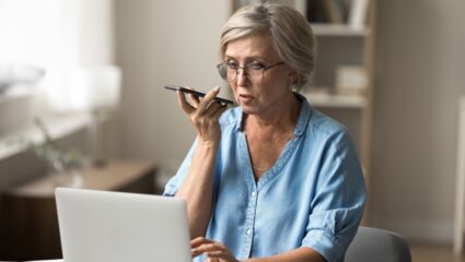 Vrouw met bril en kort grijs haar achter laptop met telefoon in haar hand