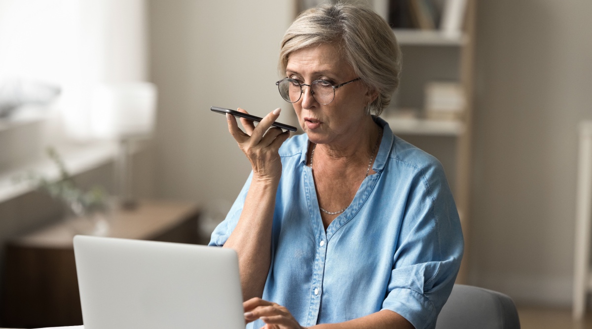Vrouw met bril en kort grijs haar achter laptop met telefoon in haar hand