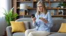 online-fraude-shutterstock_2442829539-1200×667 Een vrouw zit met haar telefoon en bankpas in de hand, in het kader van online fraude.