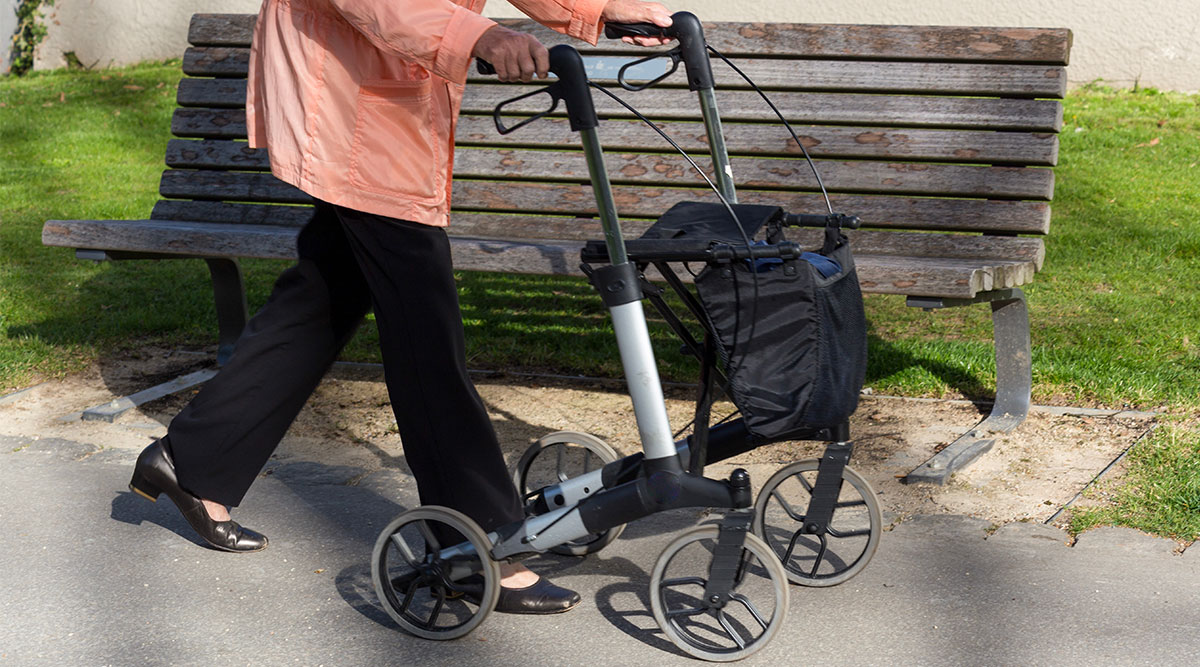 Een vrouw loopt met een rollator langs een bankje.