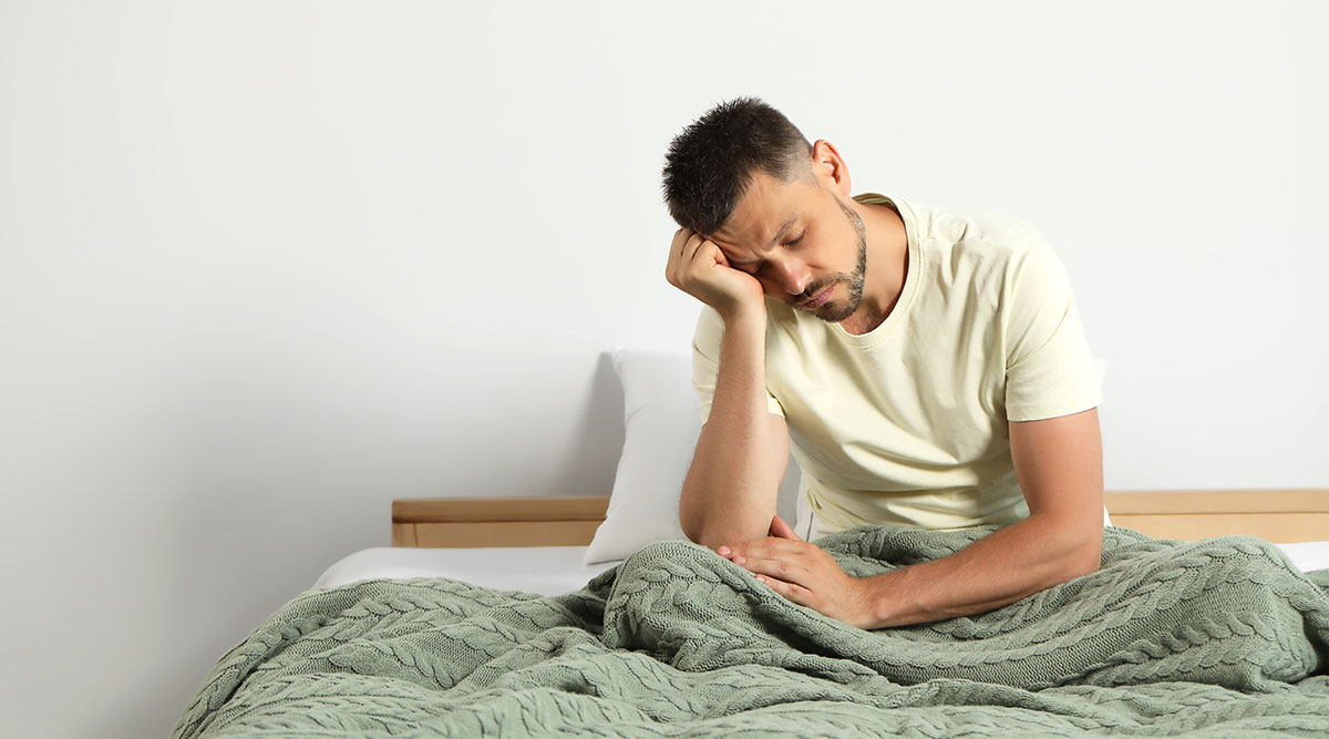 Een man met slaaptekort zit op bed en oogt vermoeid.