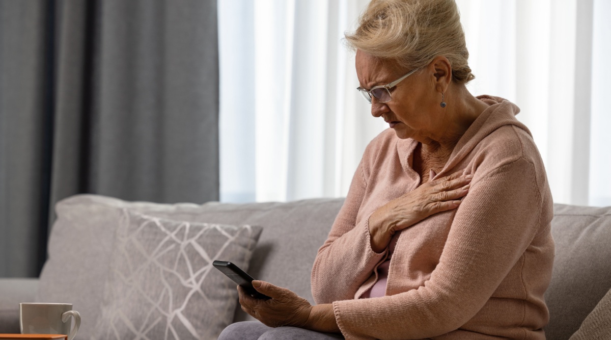 Vrouw met bril op en met smarthphone in haar hand op de bank en kijkt geschokt